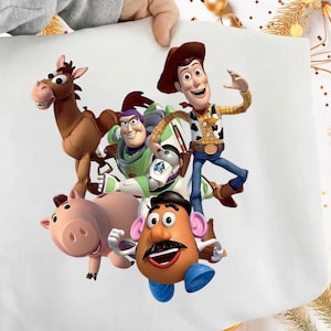May include: A white fabric featuring a colorful illustration of characters from the animated film Toy Story. The characters include Woody, Buzz Lightyear, Mr. Potato Head, Hamm, and Bullseye. The image is held by a person.