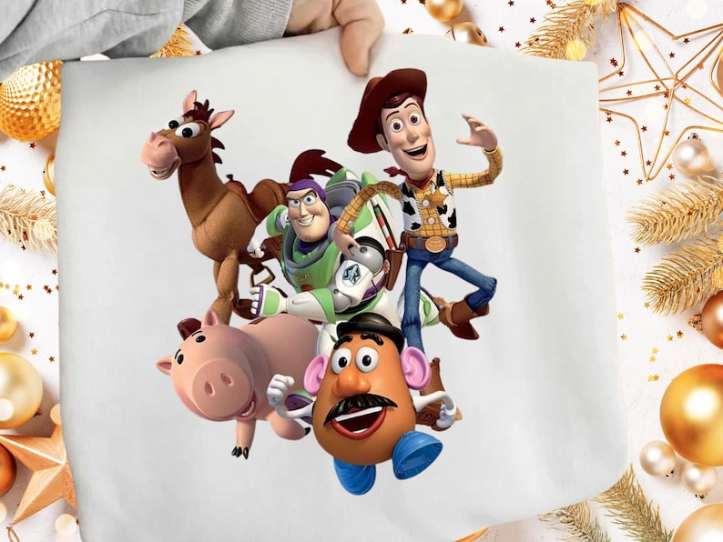 May include: A white fabric featuring a colorful illustration of characters from the animated film Toy Story. The characters include Woody, Buzz Lightyear, Mr. Potato Head, Hamm, and Bullseye. The image is held by a person.