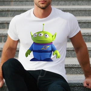 May include: White t-shirt featuring a cartoon alien character with three eyes, green skin, and blue clothing. The alien has a purple collar and a yellow emblem. The shirt is made of a soft material.
