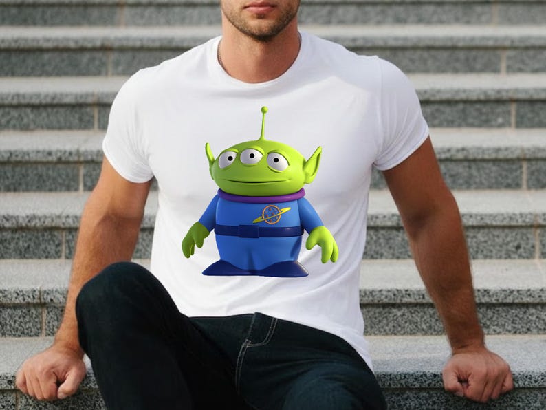 May include: White t-shirt featuring a cartoon alien character with three eyes, green skin, and blue clothing. The alien has a purple collar and a yellow emblem. The shirt is made of a soft material.