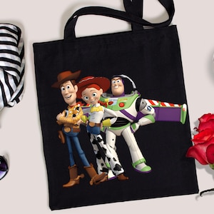 May include: Black tote bag featuring Woody, Jessie, and Buzz Lightyear from Toy Story. The bag has black handles and a colorful graphic of the animated characters. A stylish accessory for fans of the movie.
