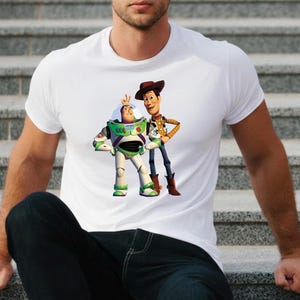 May include: White t-shirt featuring a graphic of Buzz Lightyear and Woody from Toy Story. Buzz Lightyear is in a green and white spacesuit, and Woody is wearing a cowboy hat and vest. The shirt is a crew neck.