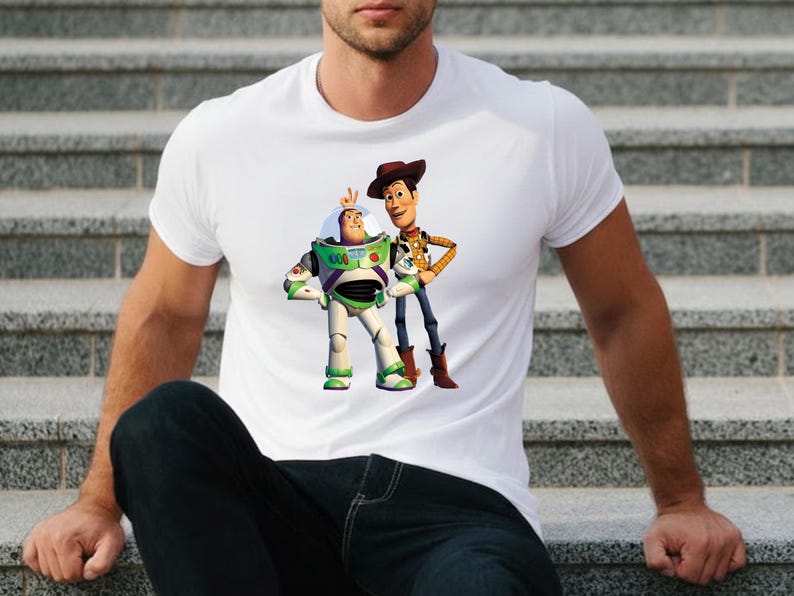 May include: White t-shirt featuring a graphic of Buzz Lightyear and Woody from Toy Story. Buzz Lightyear is in a green and white spacesuit, and Woody is wearing a cowboy hat and vest. The shirt is a crew neck.
