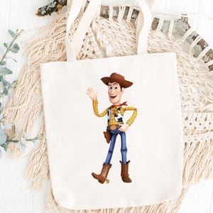 May include: A white canvas tote bag featuring a colorful illustration of the character Woody from the movie Toy Story. Woody is depicted in his signature cowboy attire, waving with a friendly expression. The bag has cream-colored handles.