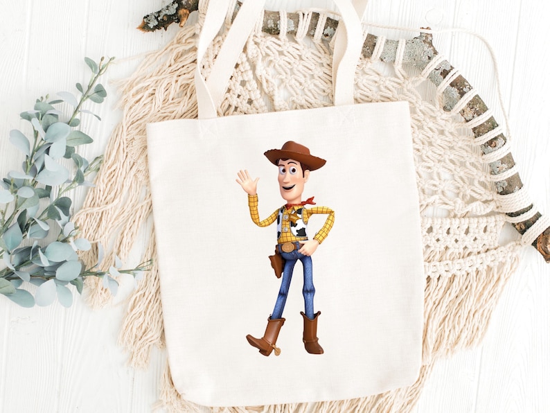May include: A white canvas tote bag featuring a colorful illustration of the character Woody from the movie Toy Story. Woody is depicted in his signature cowboy attire, waving with a friendly expression. The bag has cream-colored handles.