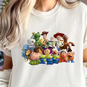 May include: Cream-colored t-shirt featuring a colorful graphic of characters from the animated movie "Toy Story." The design includes Woody, Buzz Lightyear, Jessie, and other beloved characters. The shirt has short sleeves and a crew neck.