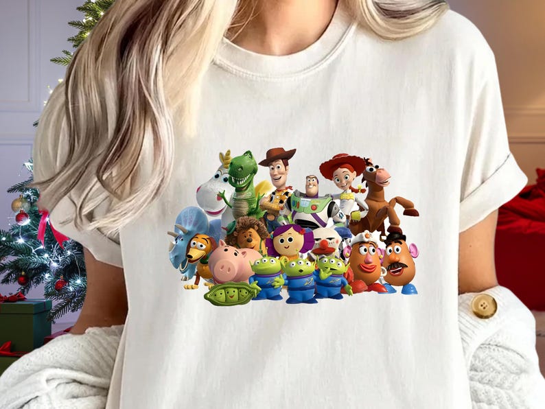 May include: Cream-colored t-shirt featuring a colorful graphic of characters from the animated movie "Toy Story." The design includes Woody, Buzz Lightyear, Jessie, and other beloved characters. The shirt has short sleeves and a crew neck.