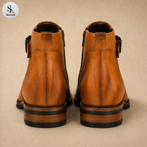 K&ouml;nnte beinhalten: Ein Paar kn&ouml;chelhohe Stiefel aus lohfarbenem Leder mit strukturierter Oberfl&auml;che, seitlichem Schnallenriemen und einem Absatz mit markanten dunklen Querstreifen. Die Stiefel sind von hinten abgebildet und zeigen die vertikale Naht am Schaft.