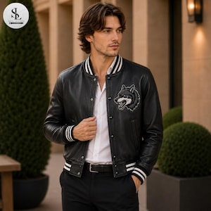 Op de afbeelding: Een zwart leren varsity-jack met een witte wolfskop-patch op de linkerborst, een witte knoopsluiting aan de voorzijde en zwart-wit gestreepte geribbelde boorden bij de kraag, mouwen en zoom, gedragen over een wit overhemd en een zwarte broek.
