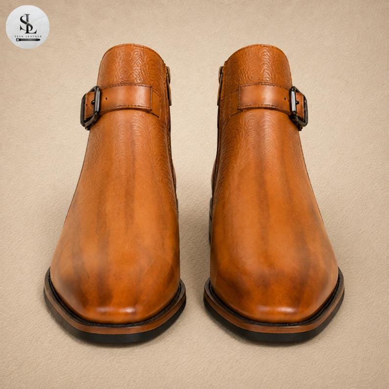 K&ouml;nnte beinhalten: Ein Paar kn&ouml;chelhohe Stiefel aus hellbraunem Leder mit strukturierter Oberfl&auml;che, spitzer Zehenpartie und einem dekorativen Riemen &uuml;ber dem Spann mit einer dunklen Metallschnalle. Die Stiefel haben eine glatte Optik mit Schattierungen und dunklem Sohlenrand.