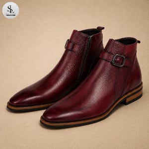 K&ouml;nnte beinhalten: Ein Paar Stiefeletten in tiefem Burgunderrot mit texturierter Lederoberfl&auml;che. Jeder Stiefel verf&uuml;gt &uuml;ber einen seitlichen Riemen mit silberfarbener Metallschnalle, einen seitlichen Rei&szlig;verschluss, eine spitze Zehenpartie und einen Holzabsatz.