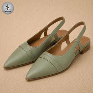 Puede incluir: Un par de bailarinas verde salvia con punta. Los zapatos tienen un detalle cosido en el empeine y un tacón bajo y apilado. Las bailarinas de cuero se muestran sobre un fondo neutro.