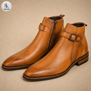 K&ouml;nnte beinhalten: Ein Paar kn&ouml;chelhohe Stiefel aus lohfarbenem Leder mit strukturierter Oberfl&auml;che, ausgestattet mit einem seitlichen Rei&szlig;verschluss an einem Stiefel und einem Zierriemen mit Metallschnalle am anderen. Die Stiefel haben eine spitze Form und einen Absatz.
