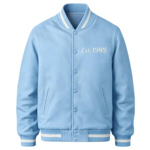 Op de afbeelding: Lichtblauwe varsity jas met witte strepen op de kraag, manchetten en zoom. De jas heeft een knoopsluiting en twee voorzakken. De tekst "Est. 1985" is op de borst geborduurd.