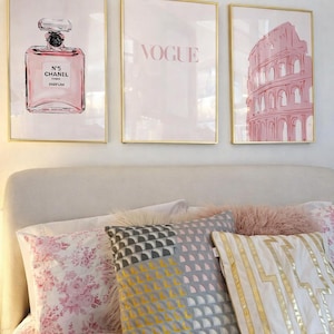 May include: Three framed art prints with gold frames hang above a bed. The prints feature a perfume bottle, the word "VOGUE", and a pink illustration of the Colosseum. The bed has floral and geometric patterned pillows.