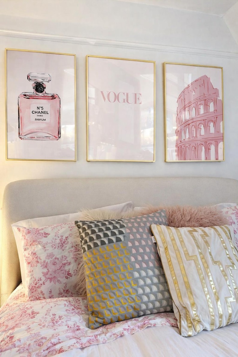 May include: Three framed art prints with gold frames hang above a bed. The prints feature a perfume bottle, the word "VOGUE", and a pink illustration of the Colosseum. The bed has floral and geometric patterned pillows.