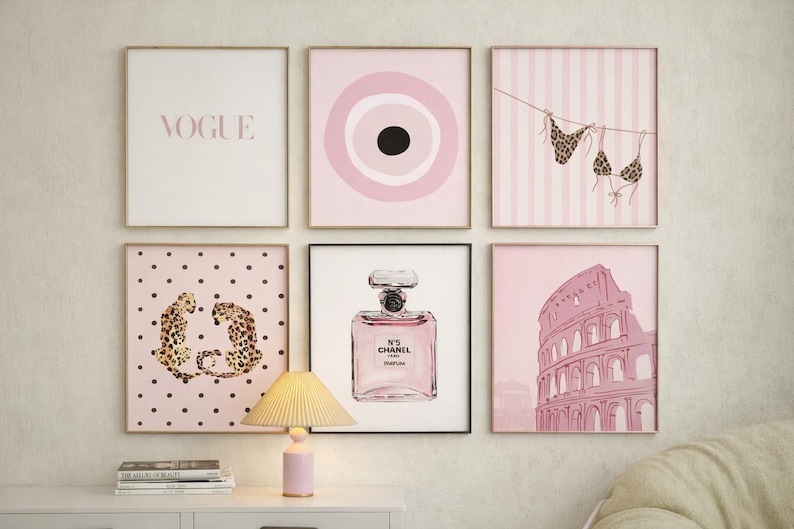 May include: A collection of framed wall art featuring a pink and gold color scheme. The art includes a Vogue print, an eye design, a bikini, leopard print, a perfume bottle, and the Colosseum. A pink lamp sits on a white table.