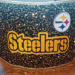 Può includere: Un contenitore nero e dorato con paillettes con la scritta "Steelers" in giallo e nero. Il logo degli Steelers è sul lato. Il contenitore è cilindrico e poggia su una superficie di legno.