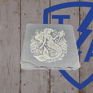 May include: A gray, square slate coaster featuring a cream-colored mermaid design. The mermaid is surrounded by decorative floral and wave patterns. The coaster is set against a wooden background with a blue logo in the upper right corner.