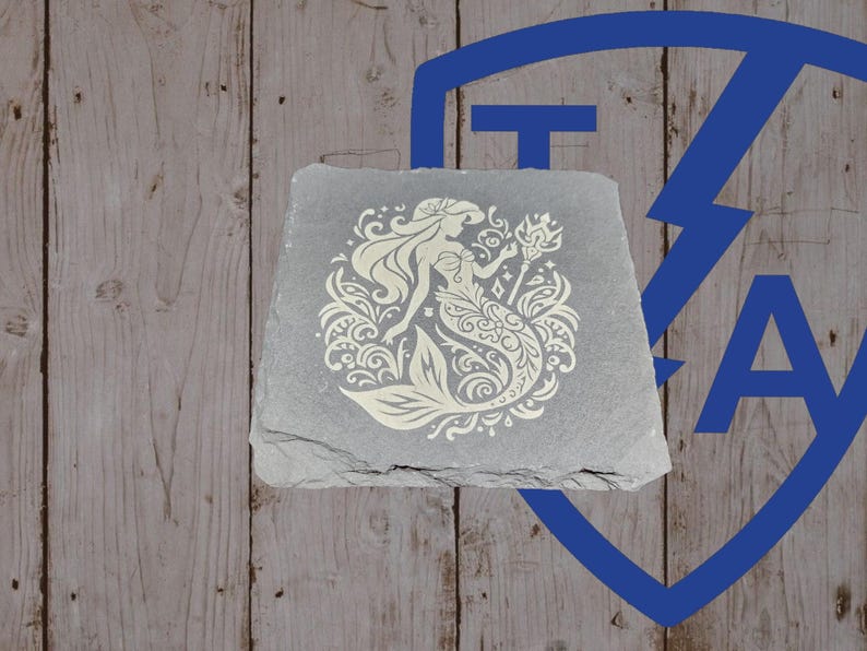 May include: A gray, square slate coaster featuring a cream-colored mermaid design. The mermaid is surrounded by decorative floral and wave patterns. The coaster is set against a wooden background with a blue logo in the upper right corner.