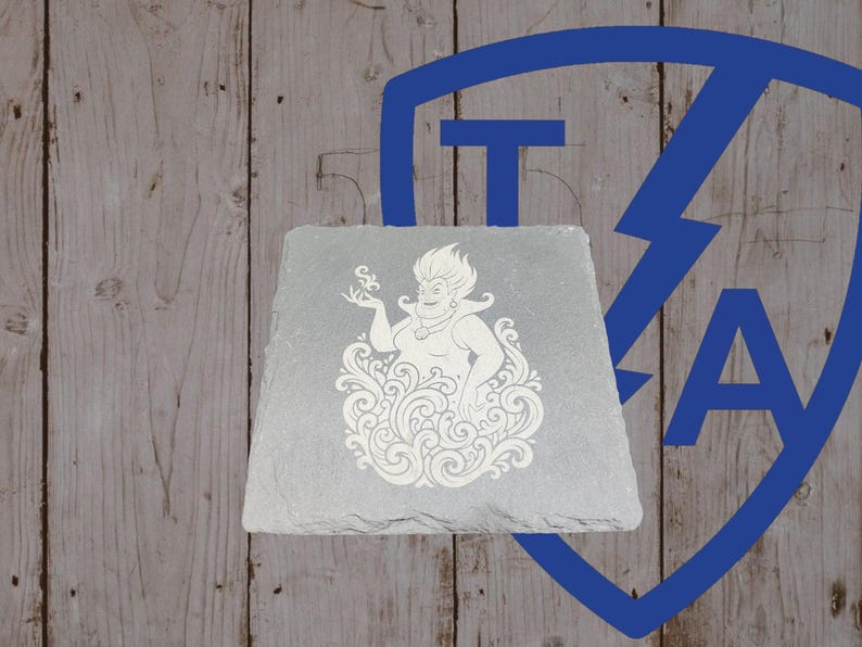 May include: A gray slate coaster featuring a white engraved image of a cartoon character. The character is surrounded by swirling lines. The background is a wooden surface with a blue logo.