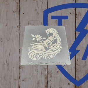 May include: A gray, square slate coaster featuring a cream-colored illustration of a woman with long flowing hair holding a flower. The design is etched into the slate. The background is a wooden surface.