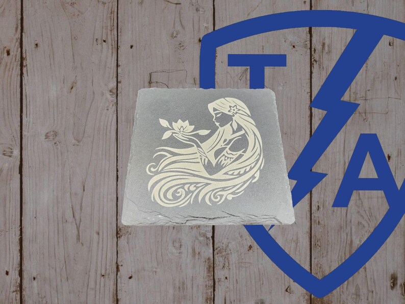 May include: A gray, square slate coaster featuring a cream-colored illustration of a woman with long flowing hair holding a flower. The design is etched into the slate. The background is a wooden surface.