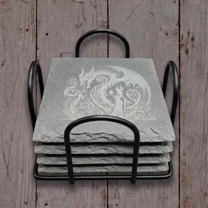 May include: A set of four gray slate coasters with a black metal holder. Each coaster features a white etched design of a dragon and a woman. The coasters are square and have a rough, natural texture.