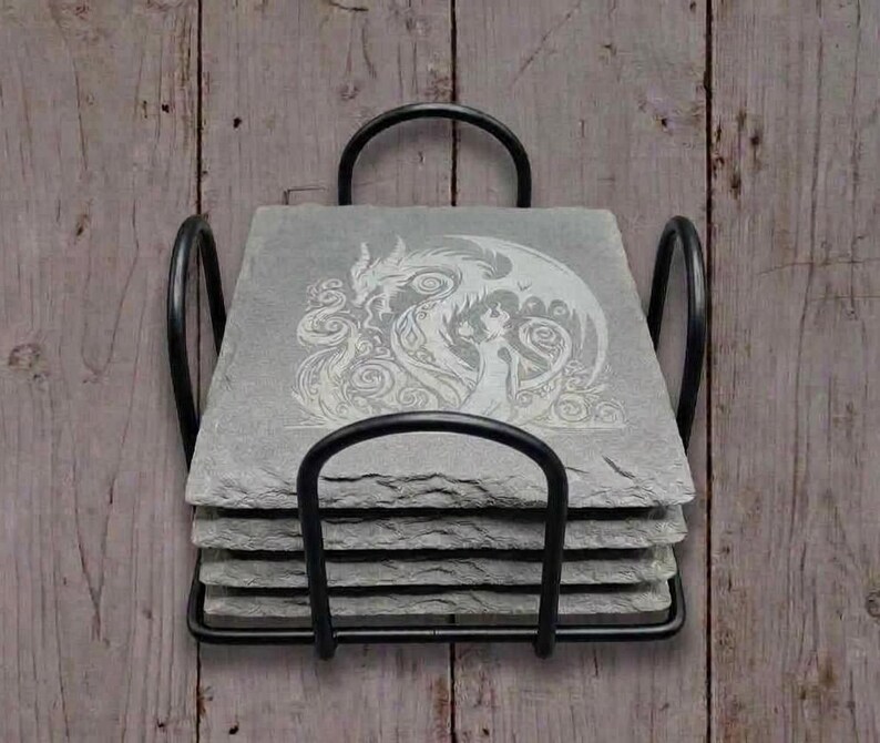 May include: A set of four gray slate coasters with a black metal holder. Each coaster features a white etched design of a dragon and a woman. The coasters are square and have a rough, natural texture.