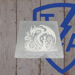May include: A gray, rectangular slate tile featuring a white, intricate design of a dragon and a woman. The dragon has large wings and is facing the woman. The woman is wearing a long dress. The background is a wooden surface.