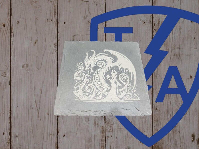 May include: A gray, rectangular slate tile featuring a white, intricate design of a dragon and a woman. The dragon has large wings and is facing the woman. The woman is wearing a long dress. The background is a wooden surface.