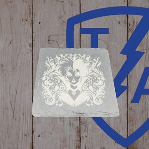 May include: A square, gray slate coaster with a white, ornate design featuring a woman's face and floral elements. The coaster is set against a wooden background with a blue graphic.