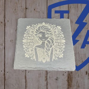 May include: A square, gray slate coaster featuring a detailed, etched image of a woman with long hair and a decorative floral design. The coaster is set against a wooden background, showcasing its natural texture and color.