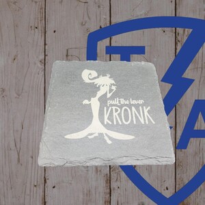 May include: A gray slate coaster with a white silhouette of a character and the text "pull the lever KRONK". The coaster has a slightly irregular shape and a textured surface. The background is a wooden surface.