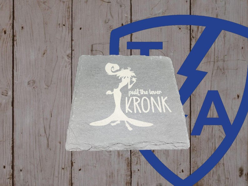 May include: A gray slate coaster with a white silhouette of a character and the text "pull the lever KRONK". The coaster has a slightly irregular shape and a textured surface. The background is a wooden surface.
