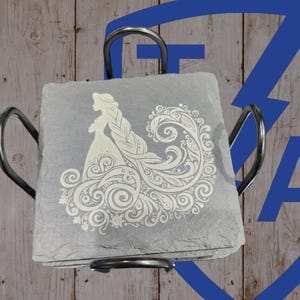 May include: A set of gray slate coasters with a white engraved design of a woman with long hair and a flowing dress. The coasters are held in a black metal holder. The background is a wooden surface.
