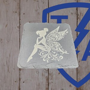 May include: A gray slate coaster featuring a white fairy design. The fairy is depicted with intricate wing details and is seated with one arm resting on her knee. The coaster has a natural, textured surface and a unique shape.