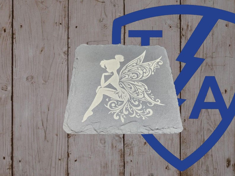 May include: A gray slate coaster featuring a white fairy design. The fairy is depicted with intricate wing details and is seated with one arm resting on her knee. The coaster has a natural, textured surface and a unique shape.