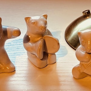 May include: Three beige ceramic bear figurines in various poses. One has arms outstretched, another holds a drum, and the third plays a stringed instrument. The figurines are on a white surface.