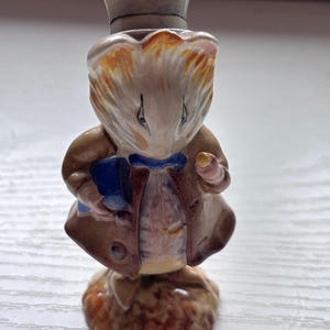 May include: A ceramic figurine of a mouse wearing a brown coat, blue bow tie, and a top hat. The mouse is holding a small blue book and a gold object. The figurine has a detailed, hand-painted design and stands on a textured base.