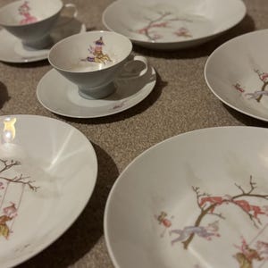May include: A collection of white porcelain teacups, saucers, and plates. The set is decorated with a playful design of figures on swings and branches in pink, gold, and purple hues.