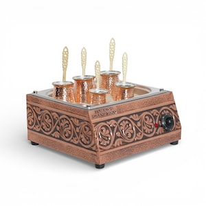 Op de afbeelding: Een koperkleurige Turkse koffiezetter met zes koperen potten en decoratieve handvatten. De basis heeft een sierlijk bloemmotief en de tekst "TURKISH GRAND SOLO". Een draaiknop bedient het verwarmingselement.