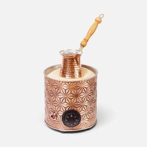 May include: A copper-colored electric coffee sand heater with a geometric star pattern on the exterior, featuring a black temperature control dial, a red indicator light, and a hammered copper coffee pot with a light-colored wooden handle.