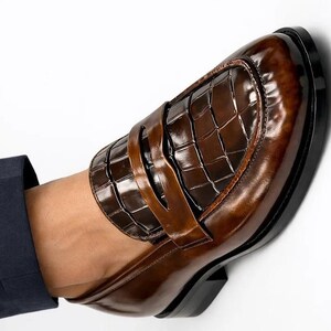 K&ouml;nnte beinhalten: Ein brauner Leder-Loafer mit gl&auml;nzender Oberfl&auml;che. Der Schuh hat ein strukturiertes, krokodilartiges Muster auf dem Blatt und einen Riemen &uuml;ber dem Spann. Die Sohle ist schwarz, vor wei&szlig;em Hintergrund.