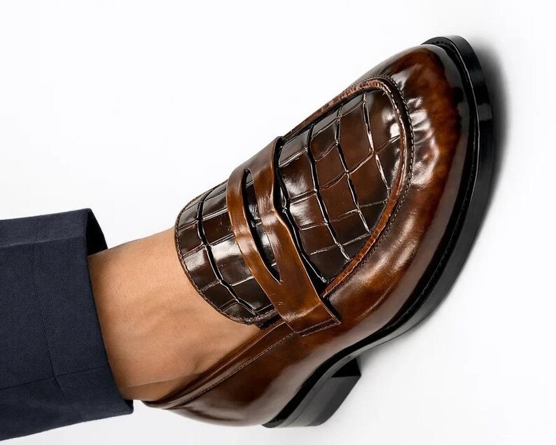 K&ouml;nnte beinhalten: Ein brauner Leder-Loafer mit gl&auml;nzender Oberfl&auml;che. Der Schuh hat ein strukturiertes, krokodilartiges Muster auf dem Blatt und einen Riemen &uuml;ber dem Spann. Die Sohle ist schwarz, vor wei&szlig;em Hintergrund.