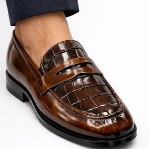 K&ouml;nnte beinhalten: Braune Leder-Penny-Loafer mit gl&auml;nzendem Finish. Die Schuhe haben einen strukturierten Vamp und eine glatte, dunkle Sohle. Das Design umfasst einen Riemen &uuml;ber dem Fu&szlig;.