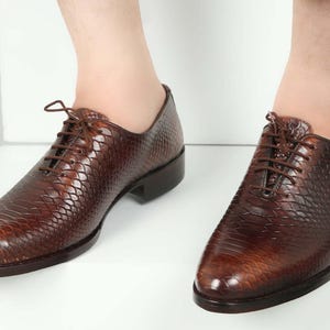 Può includere: Un paio di scarpe eleganti in pelle marrone con un motivo strutturato simile alla pelle di serpente. Le scarpe hanno un design classico con lacci e un tacco basso. La pelle ha una finitura lucida. Adatte per occasioni formali.