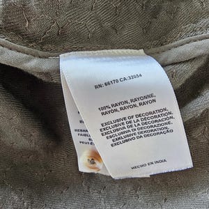 May include: Close-up of a textured olive green fabric with a white tag attached. The tag includes text such as "100% RAYON" and "EXCLUSIVE OF DECORATION". A small button is visible.