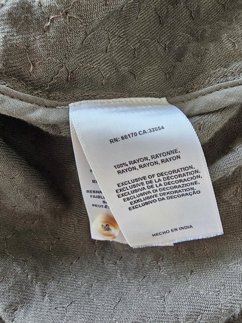 May include: Close-up of a textured olive green fabric with a white tag attached. The tag includes text such as "100% RAYON" and "EXCLUSIVE OF DECORATION". A small button is visible.