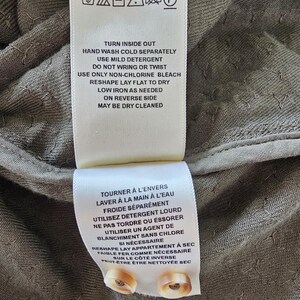 May include: Close-up of two white fabric tags with care instructions in English and French. The tags are attached to a textured, olive-green fabric. The English tag includes washing and ironing instructions. Two beige buttons are visible.